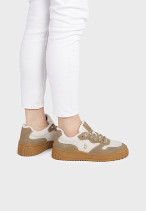Personne portant un pantalon blanc et des baskets beiges avec doublure en fourrure blanche et semelles marron, debout sur un fond gris clair.