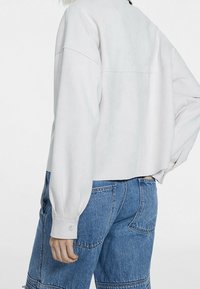 Personne portant une chemise blanche à manches longues ample, rentrée dans un jean en denim bleu taille haute, avec une main reposant près de la hanche.