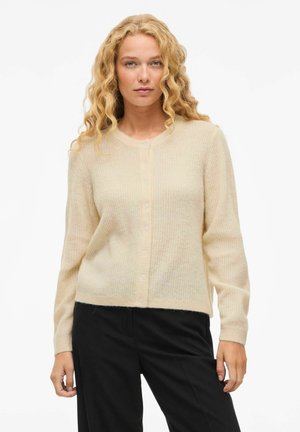 Cardigan de couleur crème en tissu doux et texturé, avec un col rond et une fermeture par boutons, présentant des détails côtelés sur le devant.