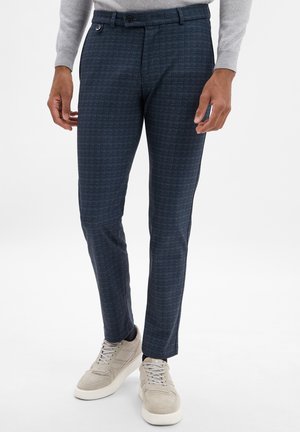 Pantaloni a scacchi blu navy con vestibilità su misura, dotati di apertura frontale piatta, tasche laterali e chiusura con bottone. Abbinati a sneaker di colore chiaro.