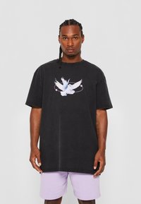 Camiseta negra oversized con un gráfico de un pájaro blanco y azul centrado en el pecho, hecha de algodón, combinada con pantalones cortos morados claros.