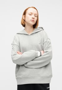 Νεαρός με κοντά κόκκινα μαλλιά, φοράει γκρι ανοιχτό φούτερ Calvin Klein και μαύρο παντελόνι, με σταυρωμένα τα χέρια, ουδέτερο φόντο.