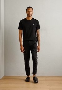 T-shirt nera a maniche corte, pantaloni cargo neri slim-fit con tasche e scarpe slip-on nere. Il modello è in piedi di fronte a uno sfondo grigio chiaro.