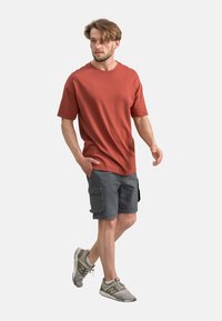 T-shirt en coton rouge, coupe ample, associé à un short cargo gris avec des poches latérales. Des chaussures de sport grises complètent la tenue.