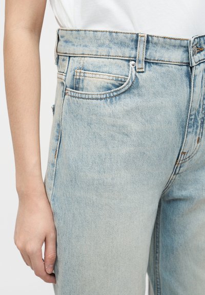 Άτομο που φοράει ανοιχτό μπλε τζιν παντελόνι ψηλής μέσης και λευκό πουκάμισο, με το αριστερό του χέρι χαλαρό δίπλα στο σώμα του.