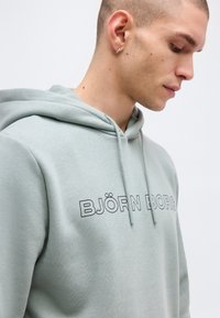 Νεαρός άνδρας φορώντας ανοιχτό γκρι φούτερ με κουκούλα με το κείμενο "BJÖRN BORG", κοιτάζει προς τα κάτω, δείχνοντας το προφίλ του και ένα ασημένιο κρίκο σκουλαρίκι στο αριστερό αυτί.