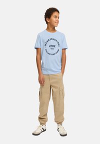 Camiseta de algodón azul claro con texto de logo circular negro; combinada con pantalones cargo beige y zapatillas blancas con acentos negros.