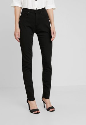 Kvinde iført tætsiddende sorte bukser, hvid skjorte med opkaldte ærmer og sorte højhælede sandaler står mod en ensfarvet baggrund.