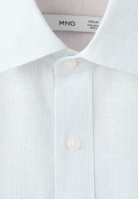 Chemise blanche texturée à boutons avec col évasé et deux boutons visibles, montrant une partie de l'étiquette de la marque MNG à l'intérieur du col.