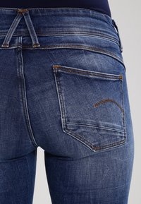 Jeans de mezclilla azul oscuro con un diseño ajustado, que presentan un detallado cosido y dos bolsillos traseros con contornos curvados. Textura suave.