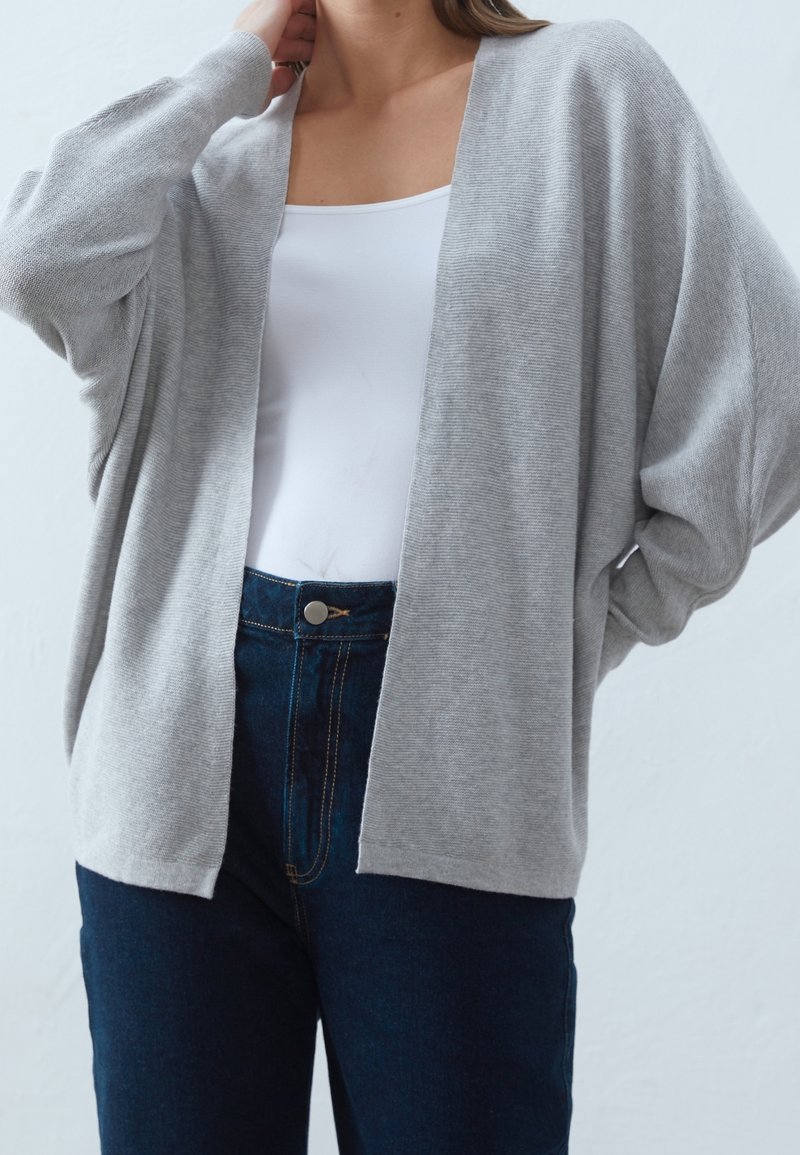Femme portant un cardigan ouvert gris clair sur un débardeur blanc et un jean bleu foncé, avec une main touchant le cou et l’autre sur la hanche.