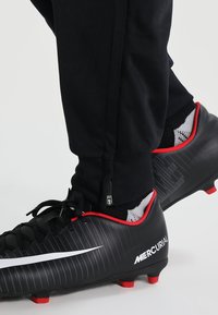 Scarpe da calcio nere con dettagli rossi e swoosh bianco. Parte superiore testurizzata, chiusura con lacci e caviglia imbottita. Pantaloni sportivi neri con cerniere.