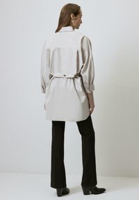 Camicia oversize grigio chiaro con vita con cintura, dettagli con bottoni e maniche lunghe. Abbinata a pantaloni a campana neri e stivaletti alla caviglia.