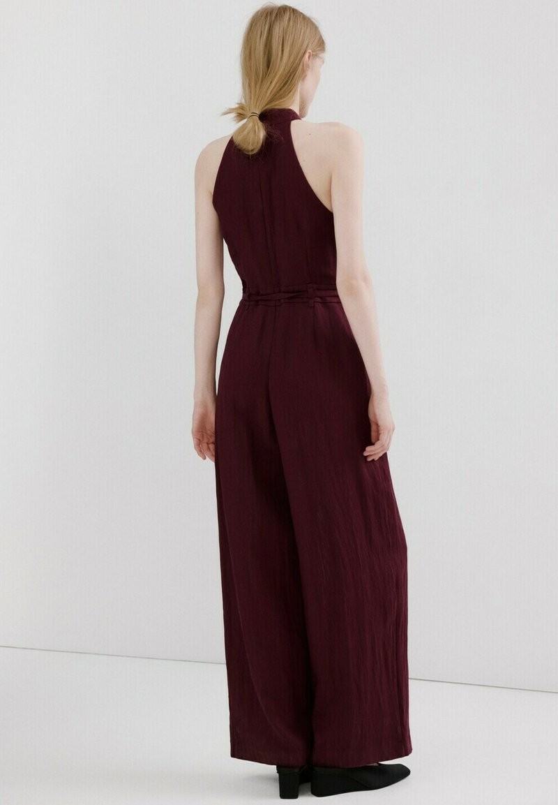 Woman standing facing a white wall, wearing a sleeveless, high-neck, dark burgundy wide-leg jumpsuit with black ankle boots.
