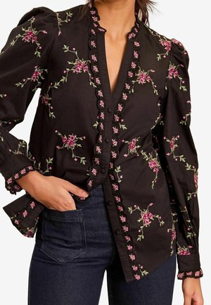Zwarte blouse met lange mouwen, voorzien van roze en groene bloemenborduursels, gegolfde randen, opgeblazen schouders en een knoopsluiting aan de voorkant, gedragen met een donkere spijkerbroek.