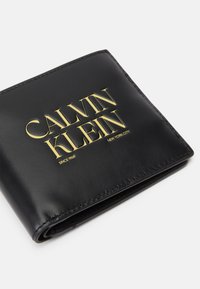 Black leather wallet with gold embossed "CALVIN KLEIN" logo, featuring smooth texture and stitching detail, folded design.