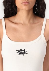 Femme portant un débardeur blanc à fines bretelles avec un logo Volcom noir en forme de soleil sur la poitrine.