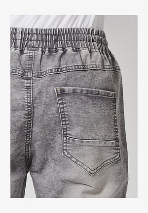 Hallus hallhidest teksapüksid veniva vöökohta, millel on taga tasku. Kanga pind on kulunud ja tekstuurne, kontrastsete õmblustega.