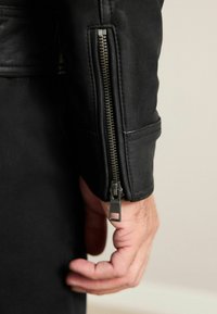 Zwarte leren jasmanchet met een metalen ritssluiting, een gladde textuur en een zakdetail vlakbij de pols, met een effen achtergrond.