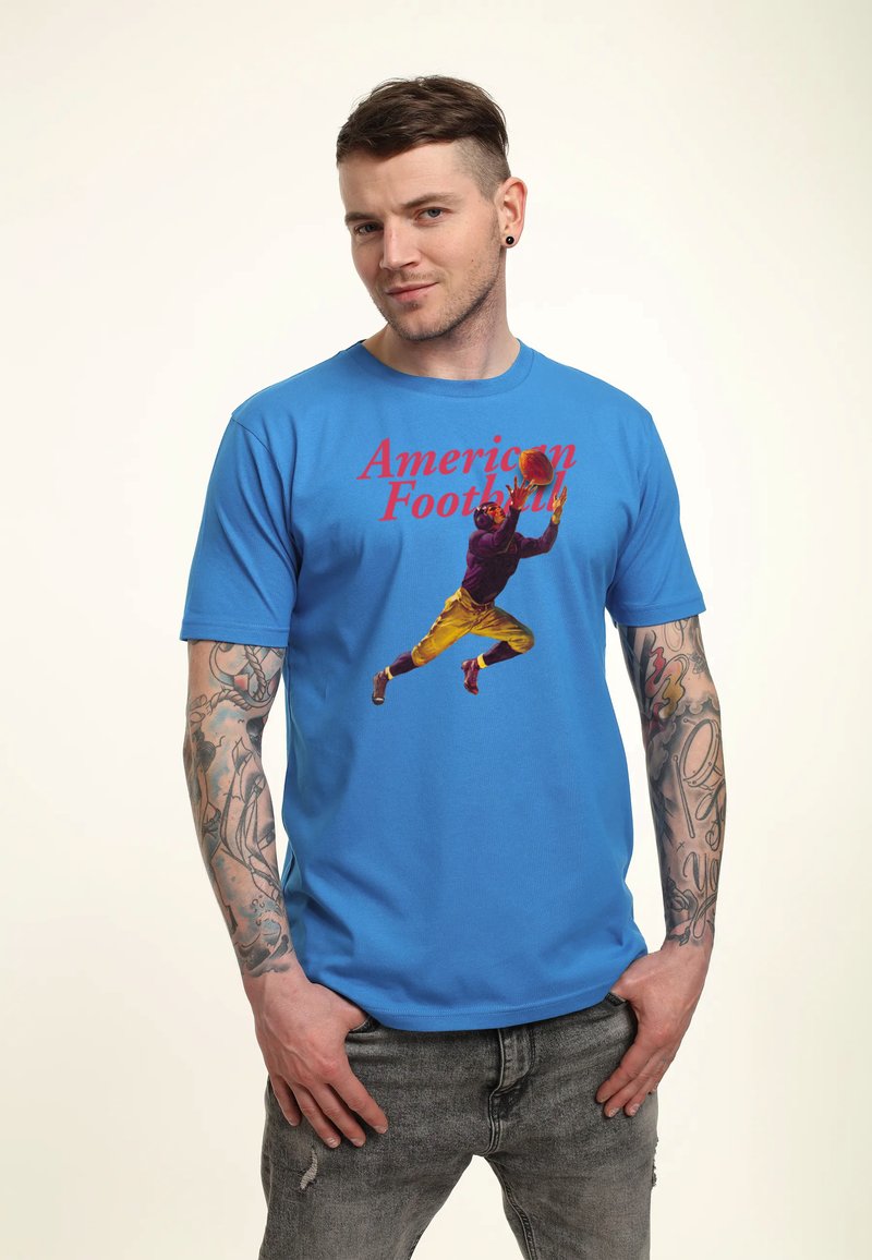 Homme tatoué portant un t-shirt bleu montrant un joueur de football américain sautant pour attraper un ballon avec le texte "Football Américain" en rouge.