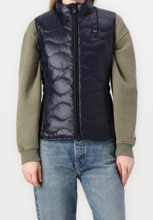 Slim fit zwart gewatteerd vest met ritssluiting aan de voorkant, gedragen over een olijfgroen shirt met lange mouwen en lichtblauwe spijkerbroek, gezien van taille tot nek.