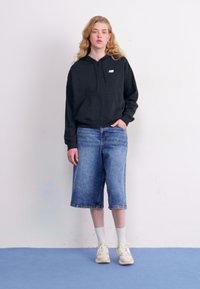 Svart zip-up hettegenser med en liten logo, kombinert med lyseblå denim Bermuda-shorts og hvite sokker, fullført med beige sneakers.