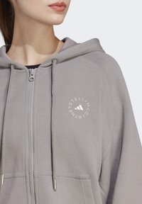 Sudadera con cremallera gris y capucha con cordones, que presenta un logo circular en el pecho. Tela suave con detalles acanalados y bolsillos delanteros.