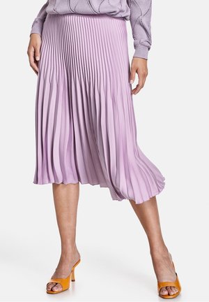 Vrouw die een lila geplooide midi-rok draagt, bijpassende trui en oranje open-teen sandalen met hak, staand met gespreide benen.