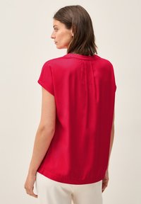 Blusa rossa a maniche corte con una texture liscia, scollatura tonda e pieghe sul retro. Taglio dritto con una vestibilità rilassata.