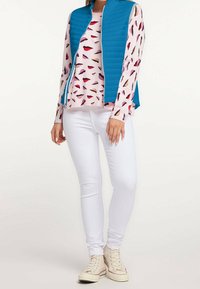 Leichter türkisfarbener Weste über einem langen, rosa Hemd mit bunten Lippenmustern, kombiniert mit weißen Skinny Jeans und beigen Sneakers.