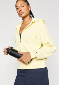 Κίτρινο φούτερ με φερμουάρ και τσέπη καγκουρό, συνδυασμένο με ναυτικό μπλε παντελόνι. Το μοντέλο κρατάει ένα μαύρο παγούρι νερού, παρουσιάζοντας μια άνετη εφαρμογή.