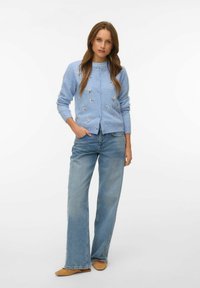 Kobieta stojąca z ręką w kieszeni, ubrana w jasnoniebieski kwiatowy kardigan, szerokie dżinsy i beżowe wsuwane buty na białym tle.