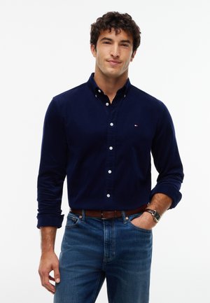 Ung mand iført en marineblå skjorte med knapper og opkørte ærmer, blå jeans, brun bælte og armbåndsur, stående med den ene hånd i lommen.