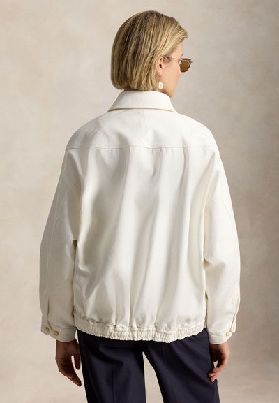 Άτομο με κοντά ξανθά μαλλιά που φορά γυαλιά ηλίου και λευκό φαρδύ μπουφάν με ελαστικό στρίφωμα, γυρισμένο προς τα πίσω σε ουδέτερο φόντο.