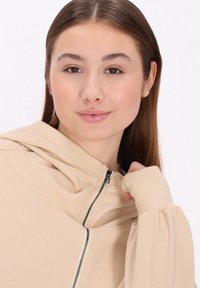 Beige színű zip-záras kapucnis felső, bordázott mandzsettákkal és puha tapintással. Ezüst cipzárral rendelkezik, laza szabású, ráncolt anyaggal.