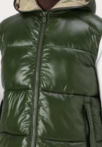 Groene puffervest van glanzend materiaal, met een ritssluiting aan de voorkant, zijzakken en een beige capuchonvoering met de tekst "DIERENVRIJ".