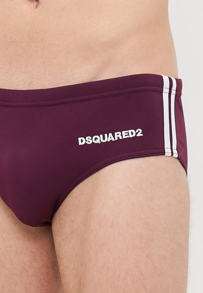 Nahaufnahme einer Person, die burgunderrote Badebriefs mit weißen Seitenstreifen und dem "DSQUARED2"-Logo am linken Hüftbereich trägt.