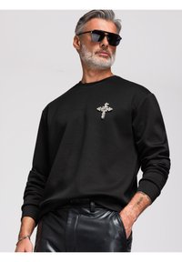 Homme aux cheveux gris et à la barbe portant des lunettes de soleil noires, un sweat-shirt noir avec une épingle croix scintillante et un pantalon en cuir noir, main dans la poche.