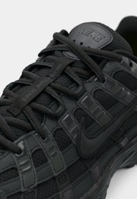 Melnas Nike sporta apavus, kas izgatavoti no sieta un gumijas materiāliem, ar teksturētu virsmas apdari un ar šņorēšanas dizainu, uz kuras atrodas Nike logo.