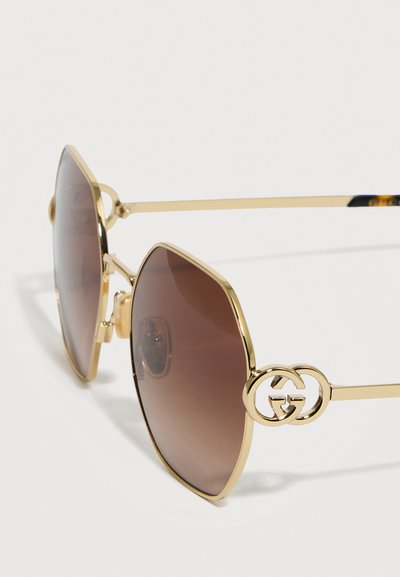 Γυαλιά ηλίου με χρυσή στεφάνη, φακούς σε καφέ διαβάθμιση και στρογγυλή λεπτομέρεια λογότυπου στους λεπτούς βραχίονες, τοποθετημένα πάνω σε λευκή επιφάνεια.