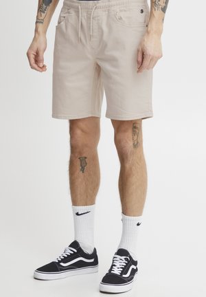 Pantalones cortos de algodón beige con cintura elástica y cordón. Dos bolsillos laterales y un ajuste casual, combinados con zapatillas negras y calcetines blancos.