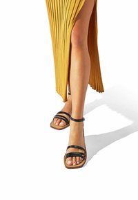Sandalias negras con tiras, de punta cuadrada, que presentan dos tiras delgadas a lo largo del pie, una sola tira en el tobillo y una suela marrón clara.