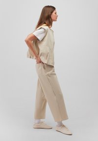 Beige ärmellose Weste über einem weißen T-Shirt, kombiniert mit lockeren beigen Hosen und hellen Sneakers, vor einem schlichten Hintergrund.