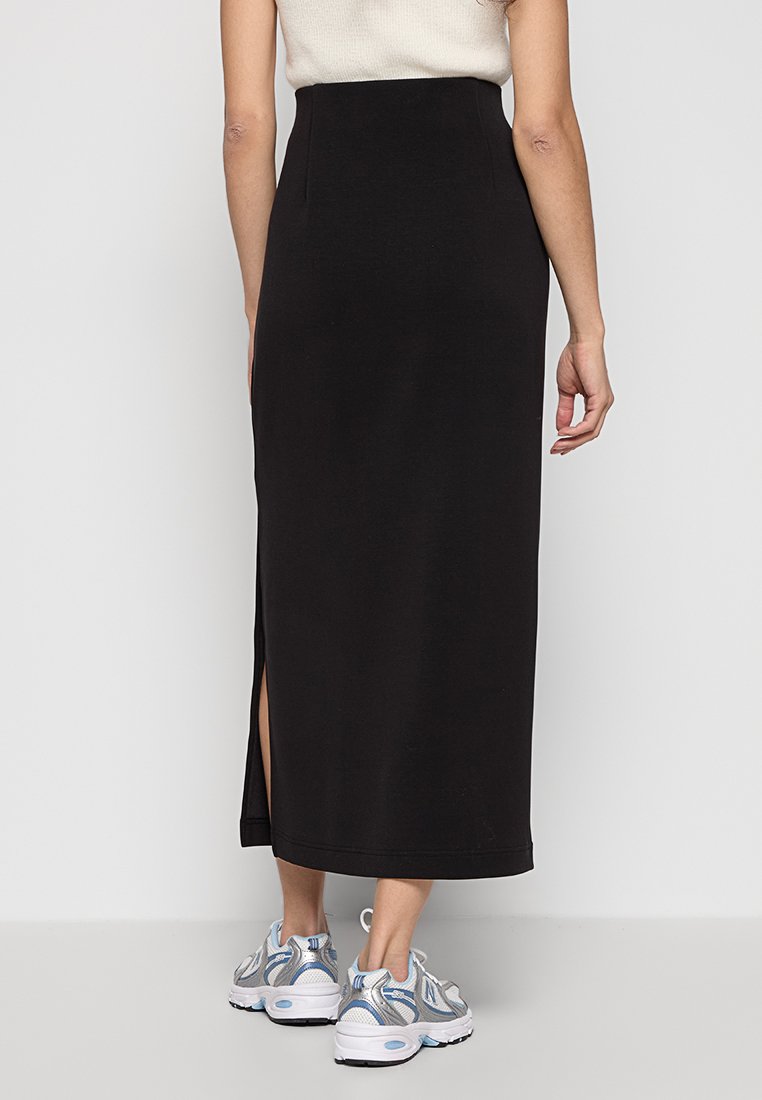 Gonna maxi nera con vita alta, caratterizzata da uno spacco laterale. Tessuto dalla texture liscia. Abbinata a sneaker azzurre e bianche.