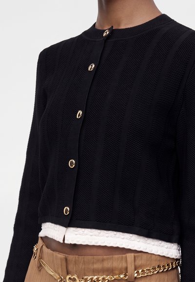 Cardigan noir texturé à boutons dorés, porté sur un top blanc texturé, assorti à un pantalon marron et une ceinture chaîne dorée.