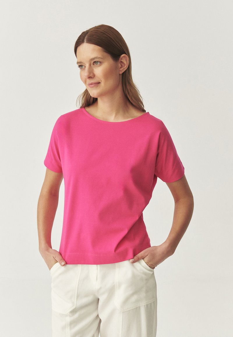 Camiseta de manga corta y cuello redondo en vibrante tela de algodón rosa, diseñada con un ajuste relajado y una textura suave. Combinada con pantalones de color claro.