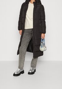 Black padded long coat with a hood, paired with a beige sweater, gray wide-leg jeans, and metallic silver platform shoes. Holds a denim handbag.