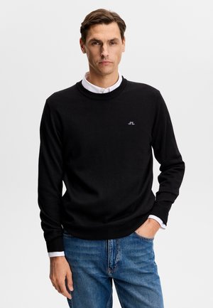 Mężczyzna w czarnym swetrze bez kołnierzyka na białej koszuli i niebieskich dżinsach, stojący z jedną ręką w kieszeni na jednolitym tle.