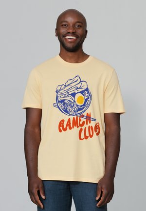 Geel katoenen t-shirt met een blauwe ramen kom grafiek met een ei en eetstokjes, bedekt met rode tekst die "RAMEN CLUB" leest.