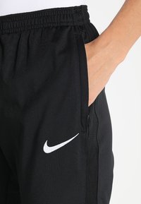 Svarta träningsbyxor av mjukt tyg med elastisk midja och sidofickor, med en vit Nike-logga på vänster ben.